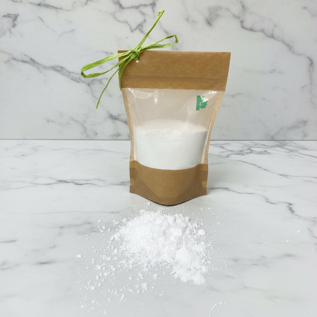 A package of dog dry shampoo with bergamot and cedarwood fragrance, accompanied by a small amount of the product spilled out onto the floor, showing the fine white powder inside. The package is brown with a green ribbon and has a label with a green leaf symbol.